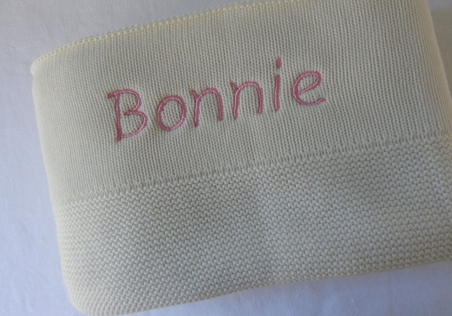 White knit baby blanket, personalised with pink thread in Italic font.