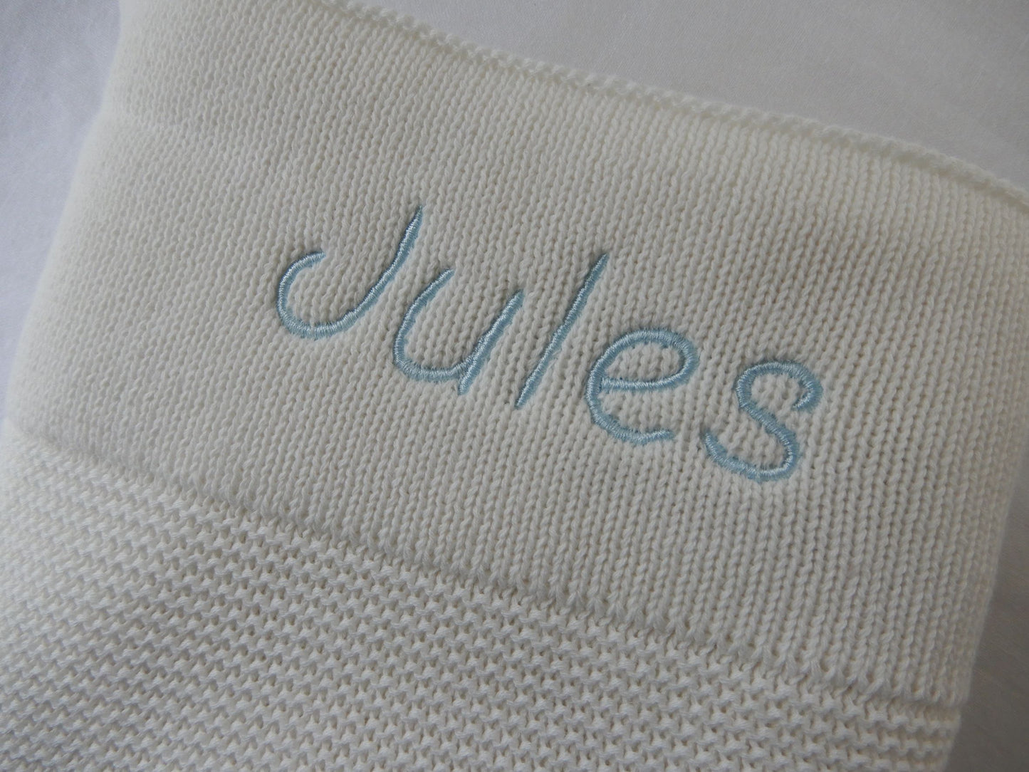 White baby knit blanket with light blue thread and italic font shown. 