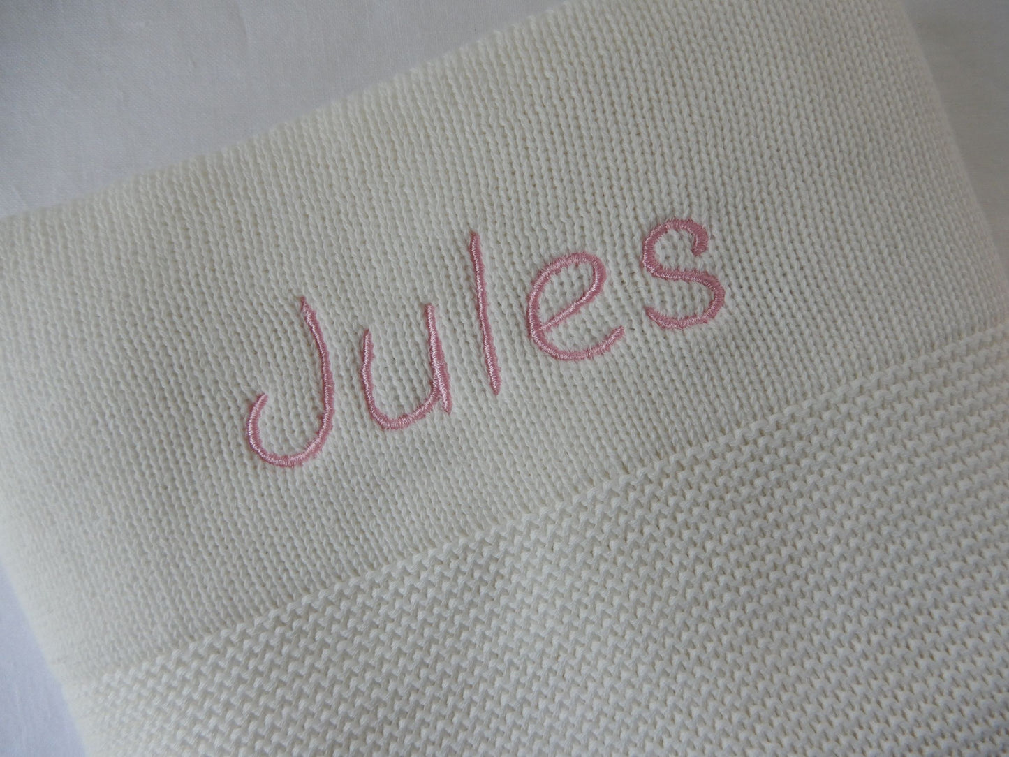 White baby knit blanket with pink thread and italic font shown. 